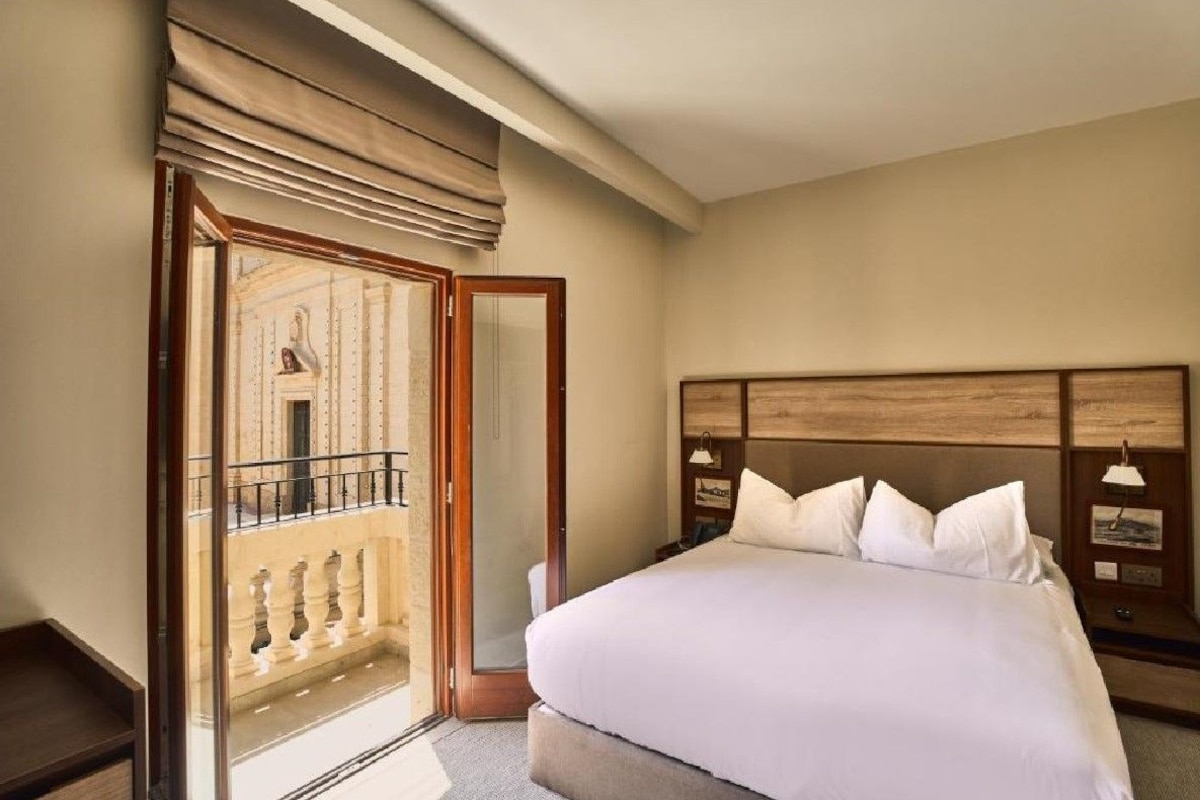 A hotel room with a large bed, a wooden headboard, a nightstand, and a balcony with a view.