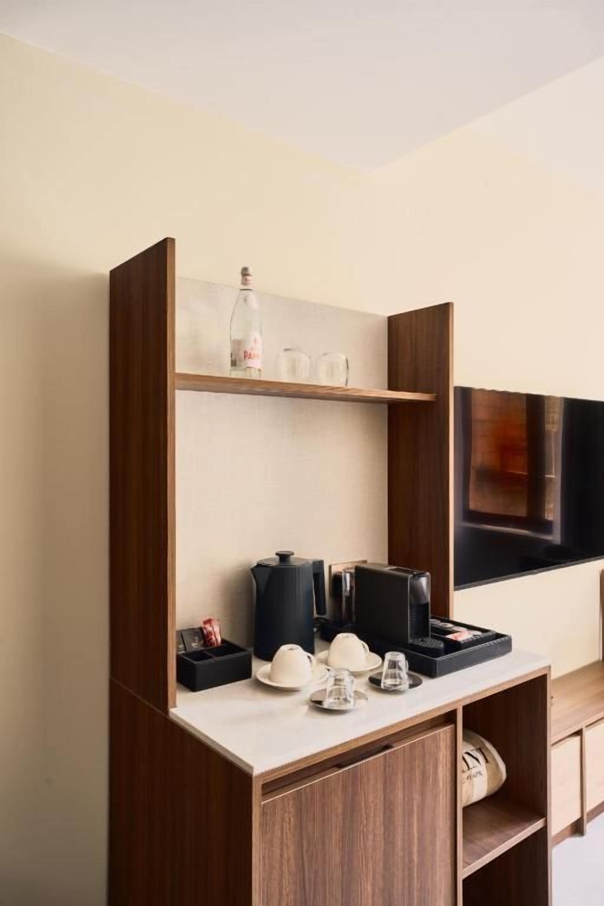 A hotel room with a wooden cabinet, a flat-screen TV mounted on the wall, and a small shelf with a bottle and glasses.