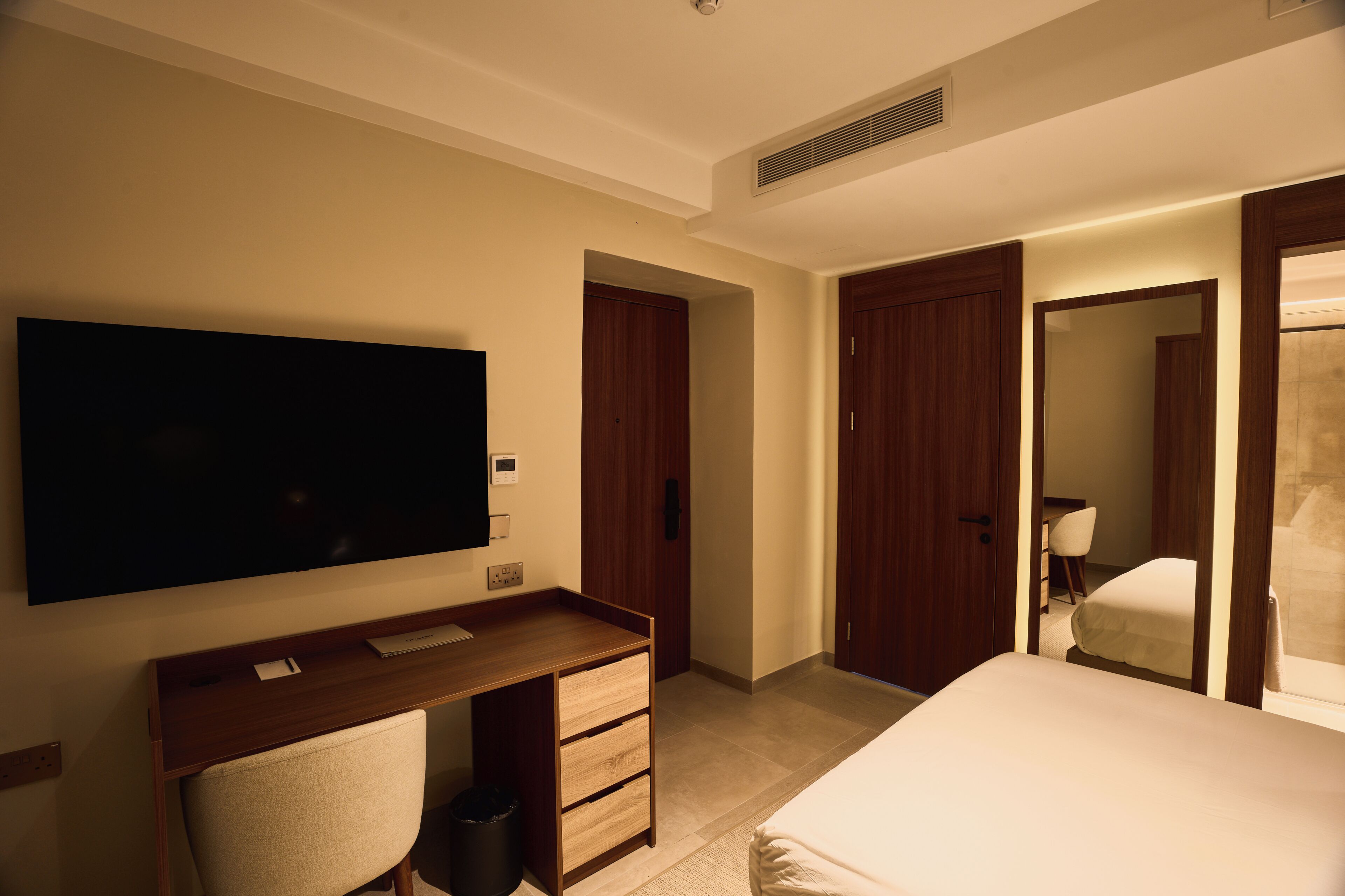 Habitación de hotel con una televisión de pantalla plana grande montada en la pared, un escritorio de madera con una silla, una cama con edredón blanco y dos puertas de madera.