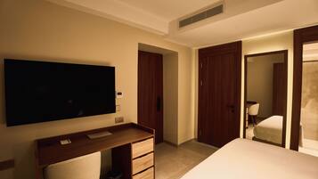 Habitación de hotel con una televisión de pantalla plana grande montada en la pared, un escritorio de madera con una silla, una cama con edredón blanco y dos puertas de madera.