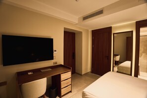 A hotel room with a large flat-screen TV mounted on the wall, a wooden desk with a chair, a bed with a white duvet, and two wooden doors. - Quaint Signature Hotel San Lawrenz (San Lawrenz)