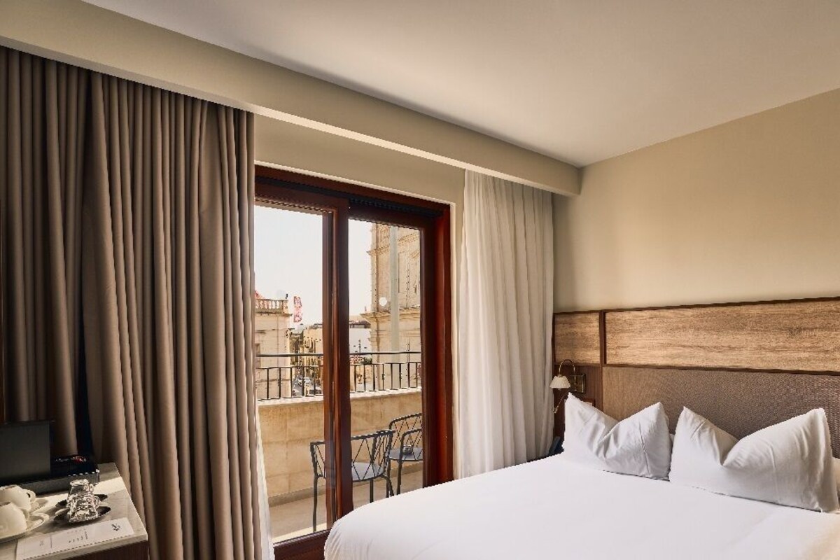 A hotel room with a bed, a desk with a telephone, and a view of a balcony with outdoor furniture.