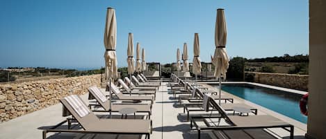 A poolside area with lounge chairs and umbrellas, a stone wall, and a clear view of the sea.