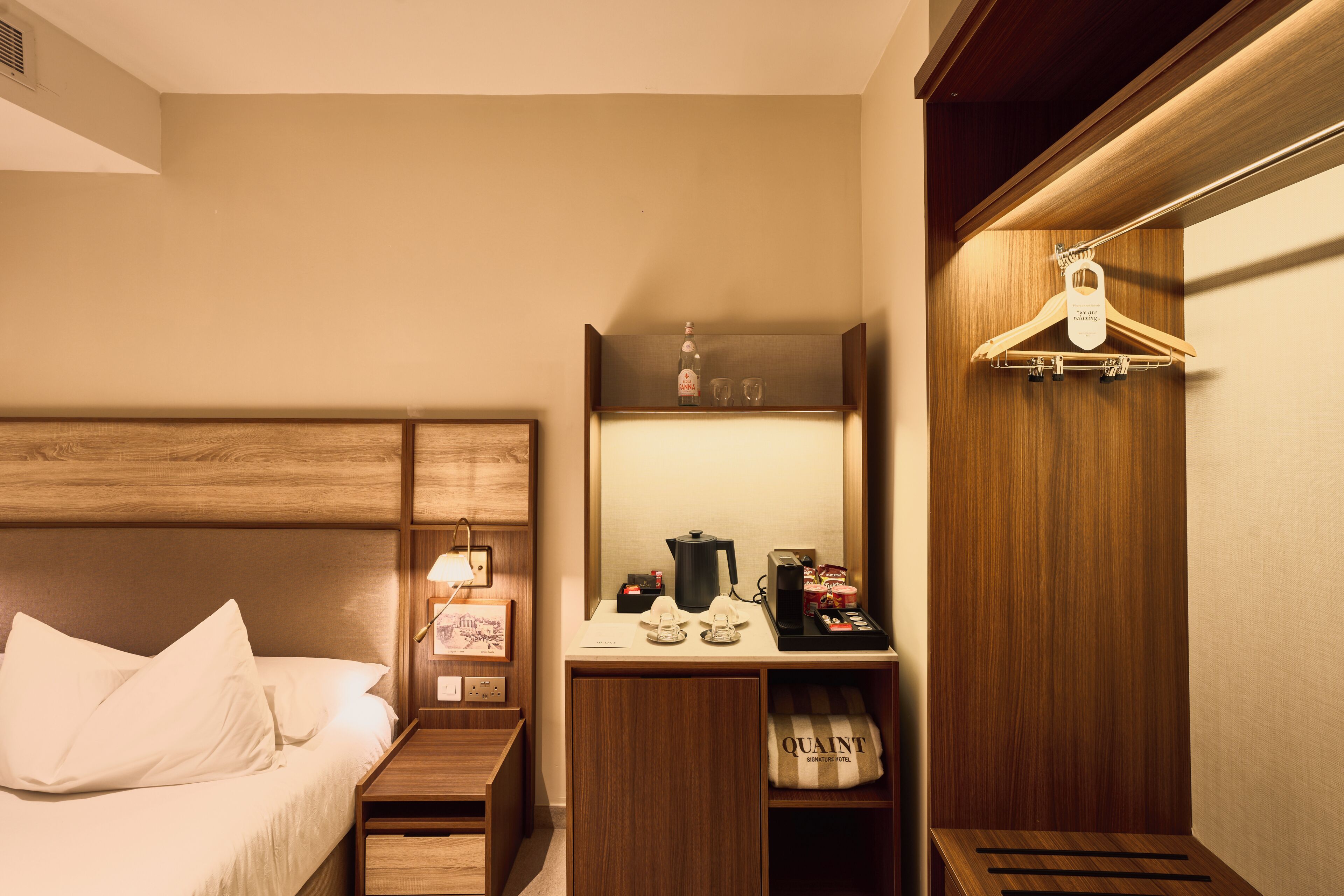 A hotel room with a bed, a nightstand, a desk with a coffee maker, a mirror, and a hanging coat rack.