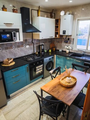 A modern kitchen with a gas stove, microwave, and built-in oven. There is a dining table with chairs and a view of the outdoors through a window. - Charming Apartment in Historic Carthage With Modern Comforts (Sidi Bou Said)
