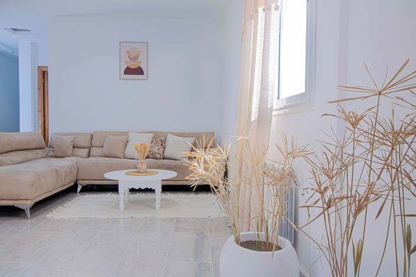 A living room with a beige sofa, a white coffee table, and a large window with natural light. - Charming Apartment in Historic Sidi Bou Said, Tunisia - Entire Rental (Sidi Bou Said)