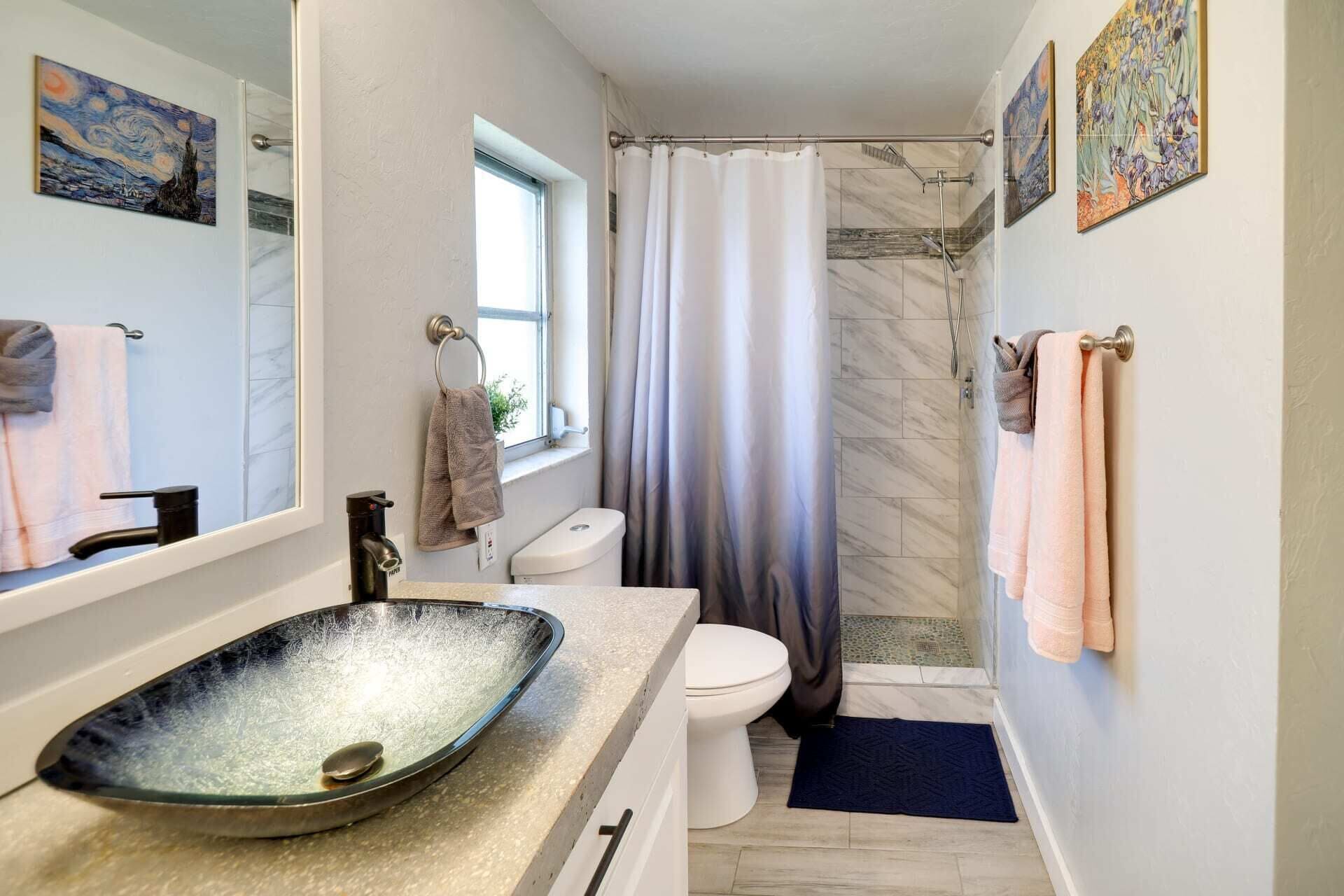 Un bagno con uno specchio grande, un lavandino dotato di una vasca originale, un water, una doccia con tenda e asciugamani su ganci.