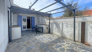 Eine überdachte Terrasse mit Steinboden, einem Tisch und einer blauen Tür, die zu einem Raum führt.