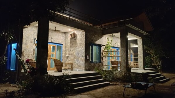 A stone building with a covered porch, steps leading to the entrance, and wicker furniture. - Camp Hornbill Corbett (Ramnagar)