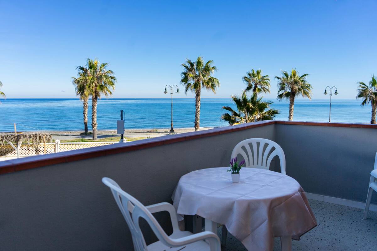A balcony with a table and chairs overlooking a beach with palm trees and the sea.