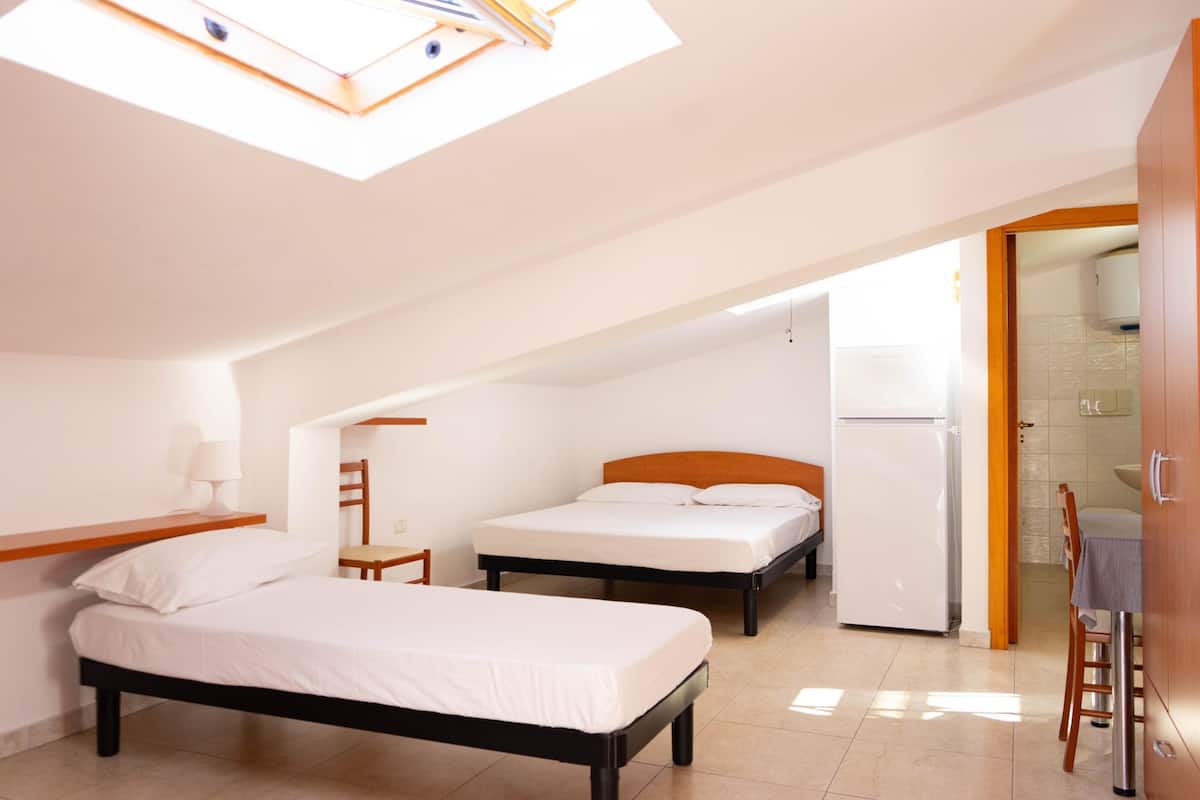 A room with two beds, a small table, a chair, a refrigerator, and a skylight.