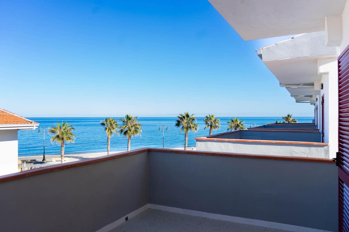 A balcony with a view of the sea, palm trees, and a building with a red awning.