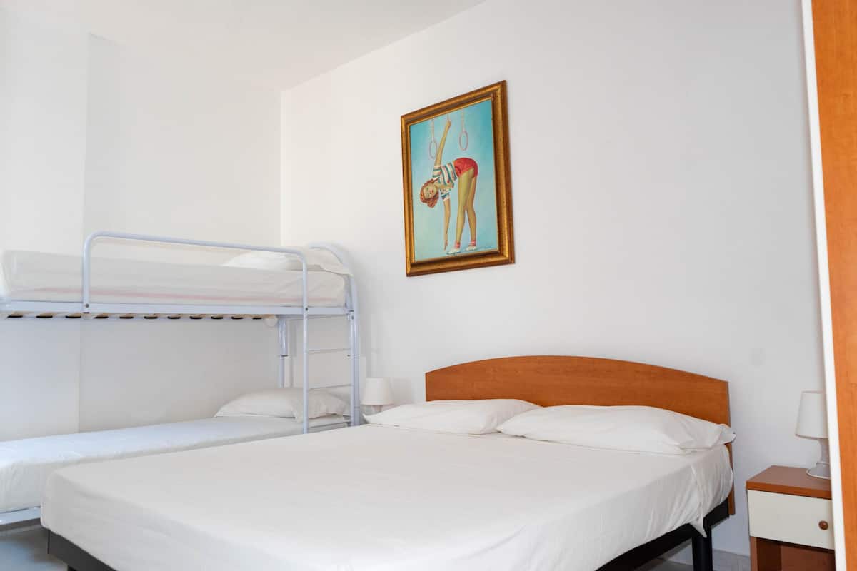 A hotel room with a bunk bed, a single bed, white linens, a wooden headboard, a framed picture on the wall, and a bedside table with a lamp.