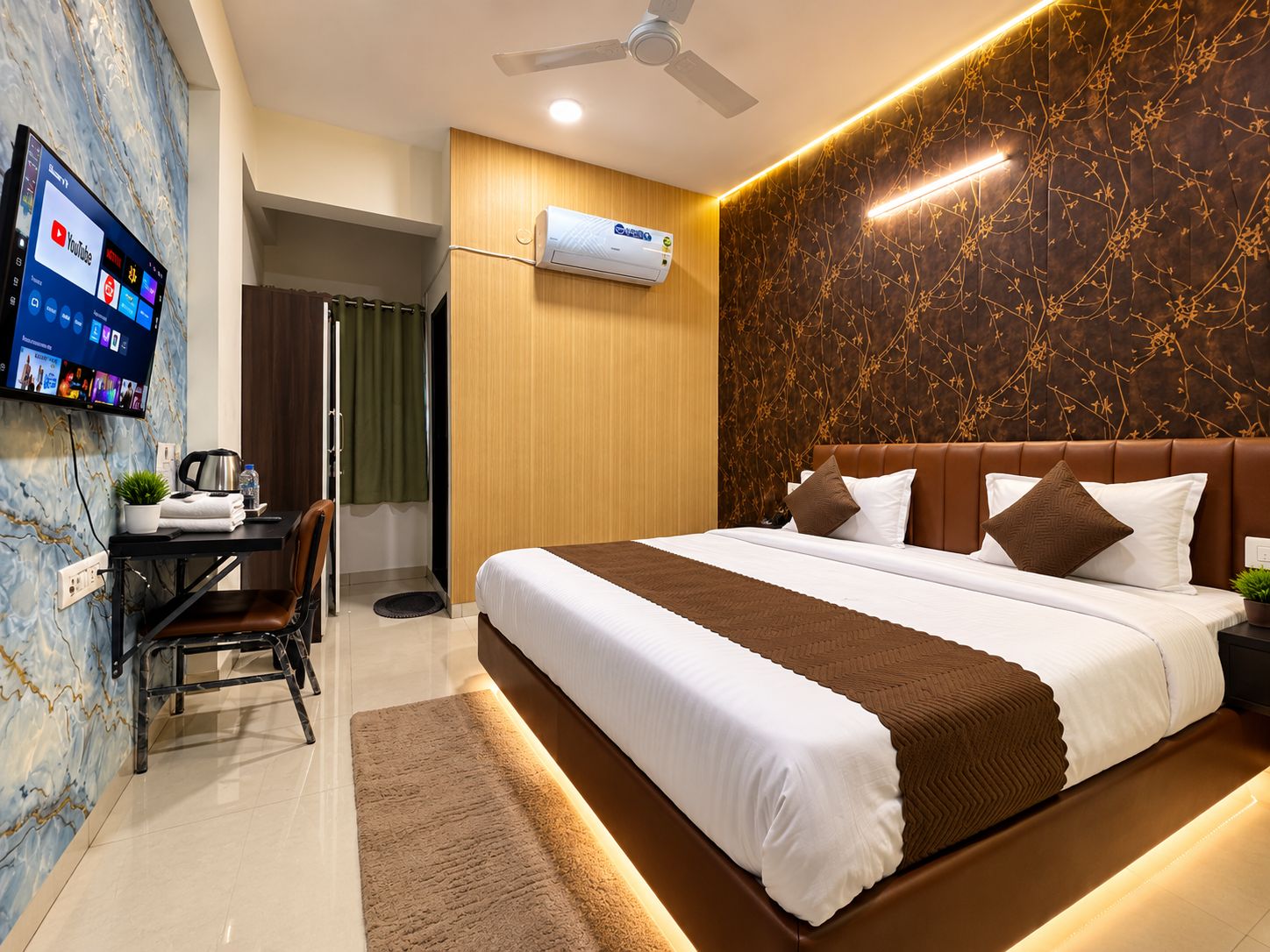 A modern hotel room with a bed, a desk with a computer, and a TV mounted on the wall.