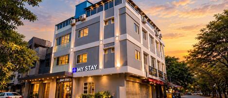 A multi-story hotel with a modern facade, a prominent 'MY STAY' sign, and a street view with parked cars and trees.