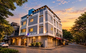 Hotel com vários andares e fachada moderna, com destaque para o letreiro 'MY STAY', vista da rua com carros estacionados e árvores.