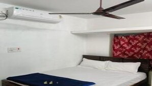 Un lit simple avec une tête de lit en bois, une literie blanche et une couverture bleue. Un ventilateur de plafond est installé au-dessus du lit, et on aperçoit une partie d’un rideau rouge à motifs.