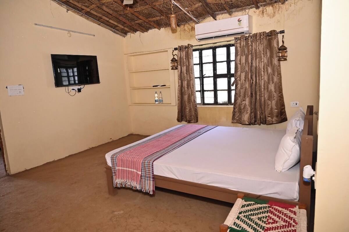 A single-bed room with a wooden frame, a flat-screen TV mounted on the wall, a window with curtains, and an air conditioning unit above it.
