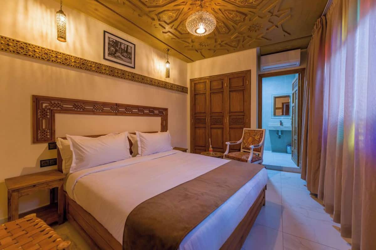 Un dormitorio con una cama grande, cabecera de madera y un techo con diseños intrincados. Hay una silla y una mesita de noche junto a la cama.