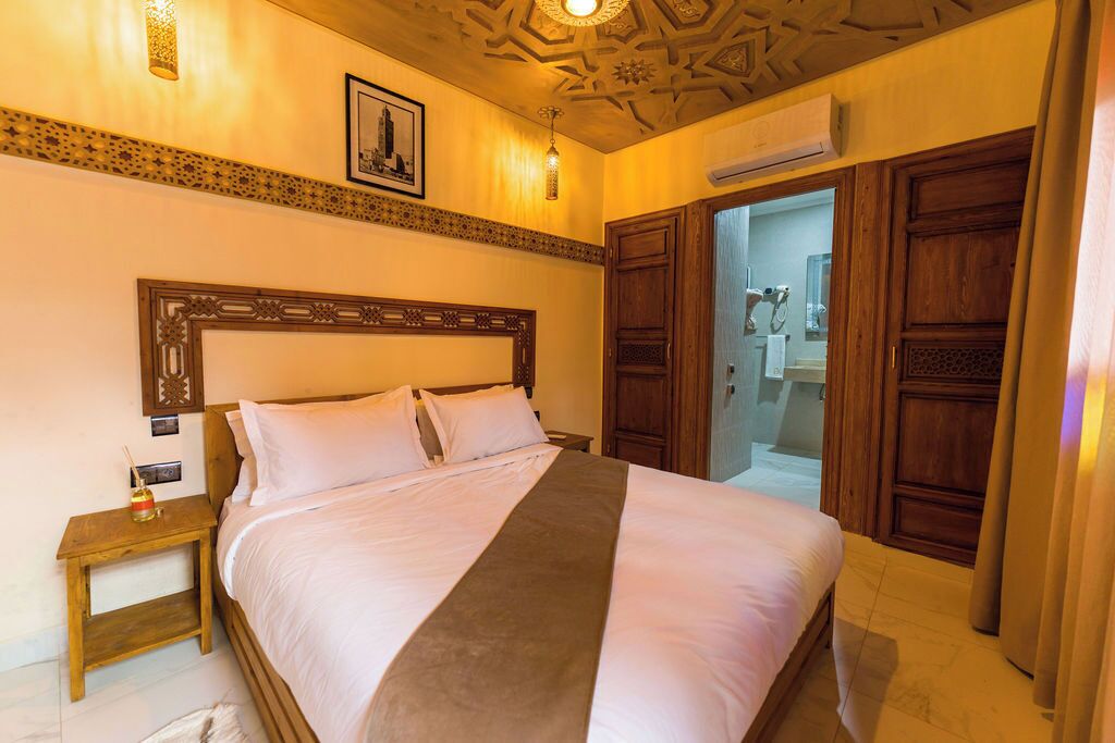 Une chambre d’hôtel avec un grand lit, une tête de lit en bois, une table de chevet avec une lampe et une porte donnant sur une salle de bain.
