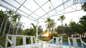An outdoor seating area with white chairs and a glass table, overlooking a pool and palm trees. - Timeless Luxury on Port Blair’s Shore (Sri Vijaya Puram)