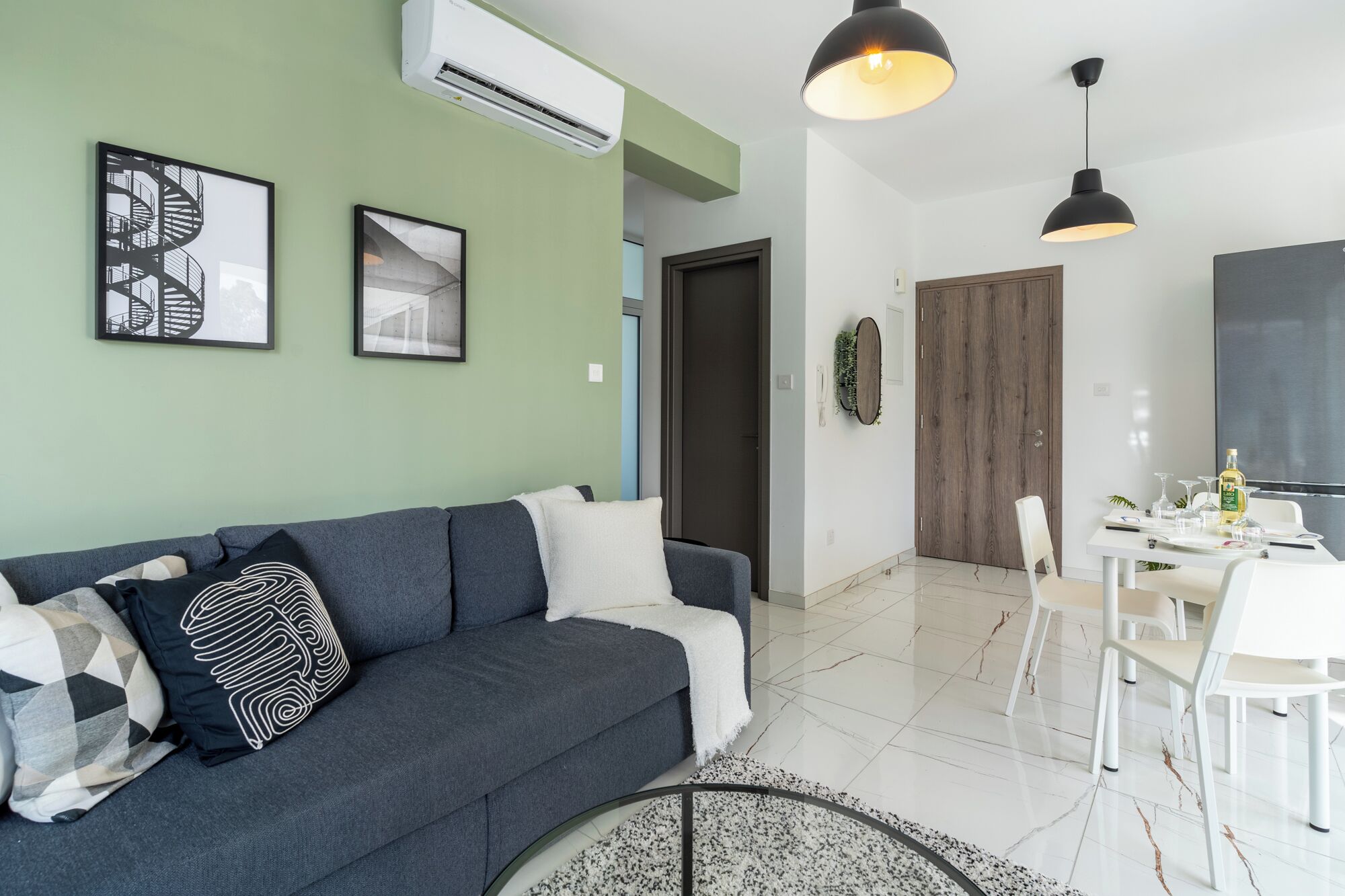 Una sala moderna con un sofá gris, dos cuadros enmarcados, una zona de comedor con una mesa y sillas blancas, y una puerta de madera.