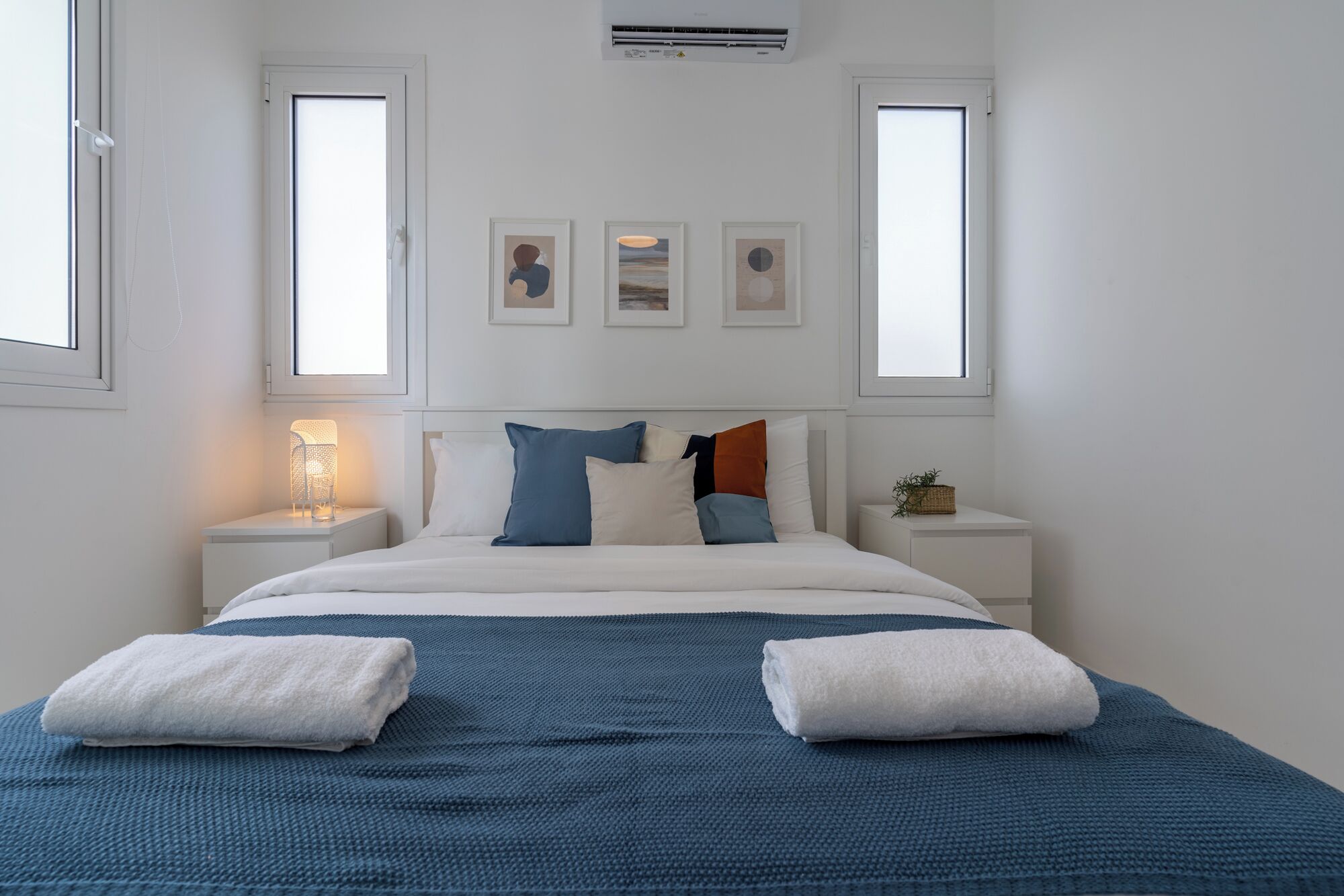 Ein Schlafzimmer mit einem Bett, zwei Nachttischen, einer Wand-Klimaanlage und drei gerahmten Kunstwerken.