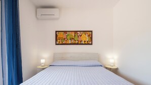 Une chambre à coucher avec un lit, deux lampes de chevet, un climatiseur fixé au mur et une œuvre d’art colorée au-dessus du lit.