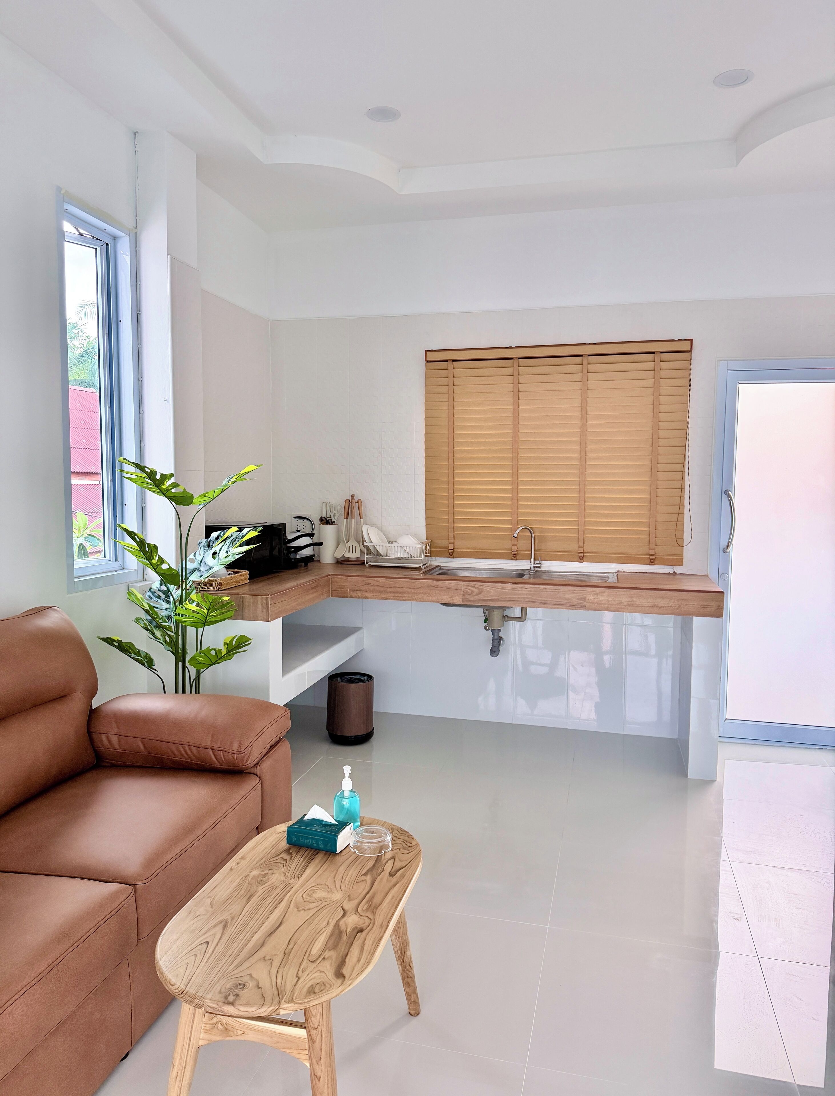 A modern kitchen with a wooden countertop, a sink, and a window with blinds. There is a brown leather sofa and a wooden coffee table with a bottle of hand sanitizer.