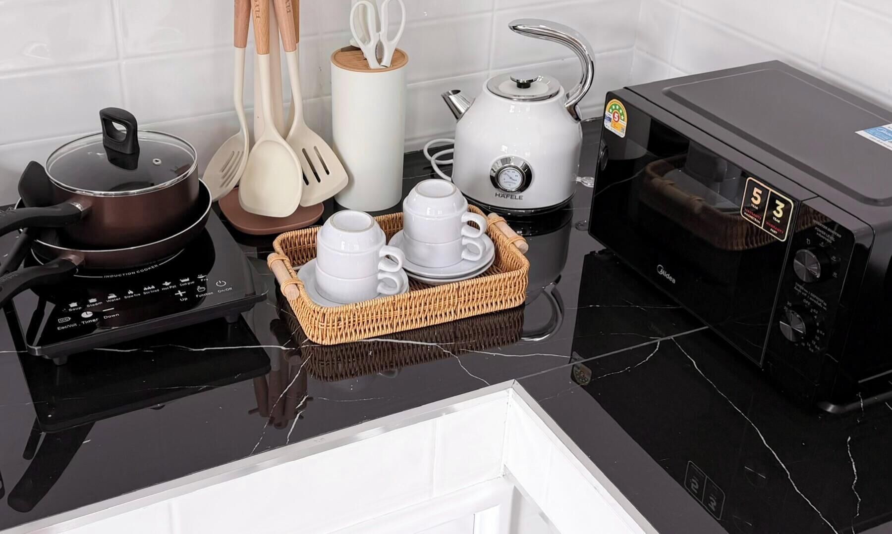 Cocina con placa de inducción, microondas, hervidor y un cesto de tazas blancas.