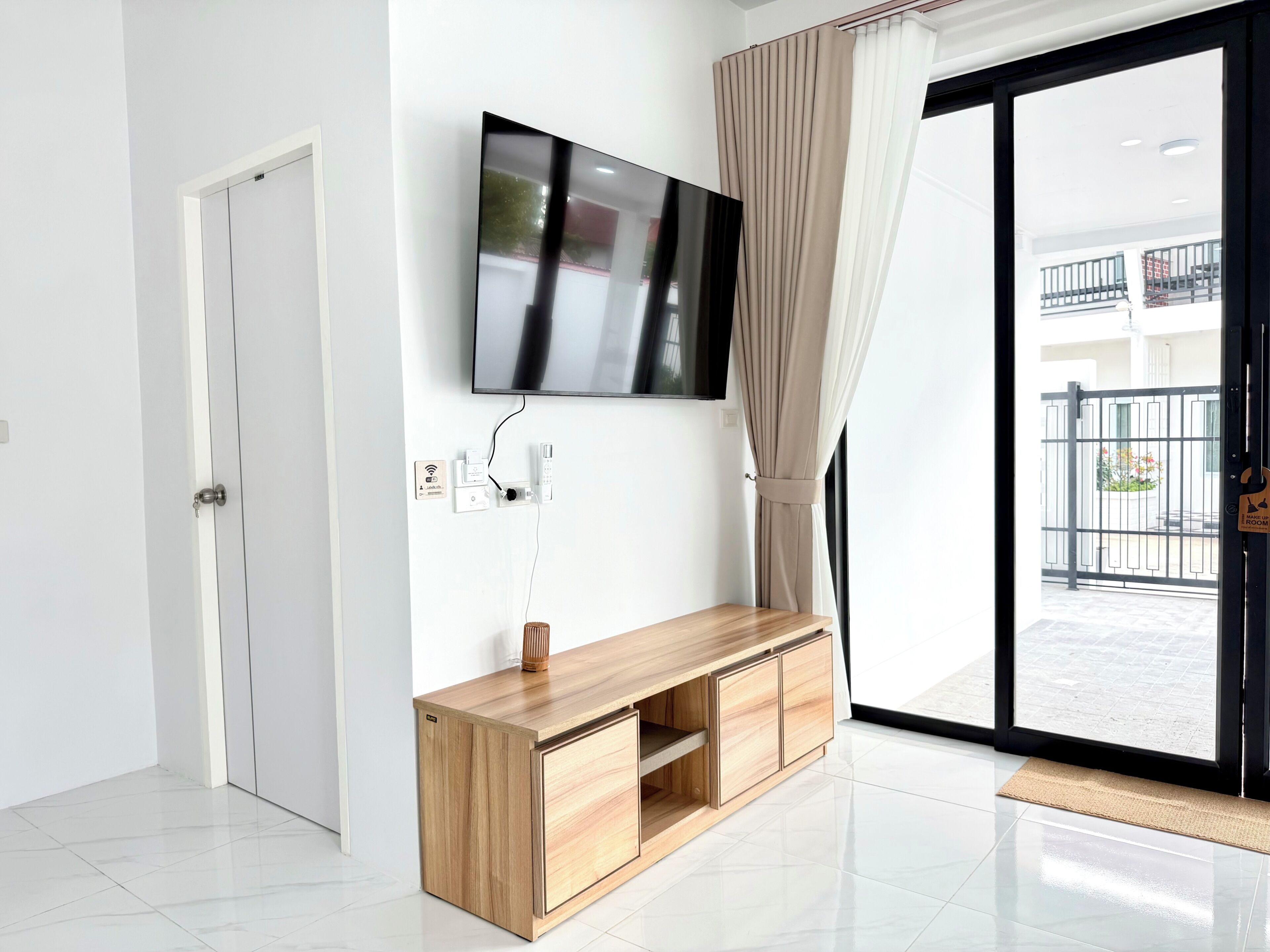A modern room with a flat-screen TV mounted on the wall, a wooden console table, a sliding glass door leading to an outdoor balcony, and a door with a handle.