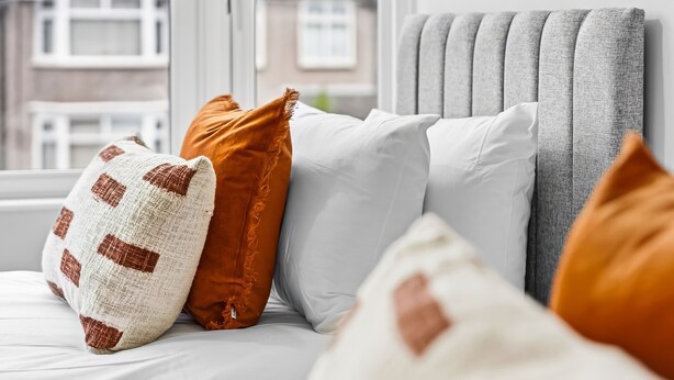 A bed with multiple pillows in various colors and patterns, a grey upholstered headboard, and a window with a view of buildings. - Cosy 3BR Home in the heart of Coventry - 1GBWiFi (Coventry)