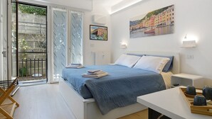Un dormitorio con cama, una mesita de noche, una silla, una ventana con vistas a un balcón y un cuadro en la pared.