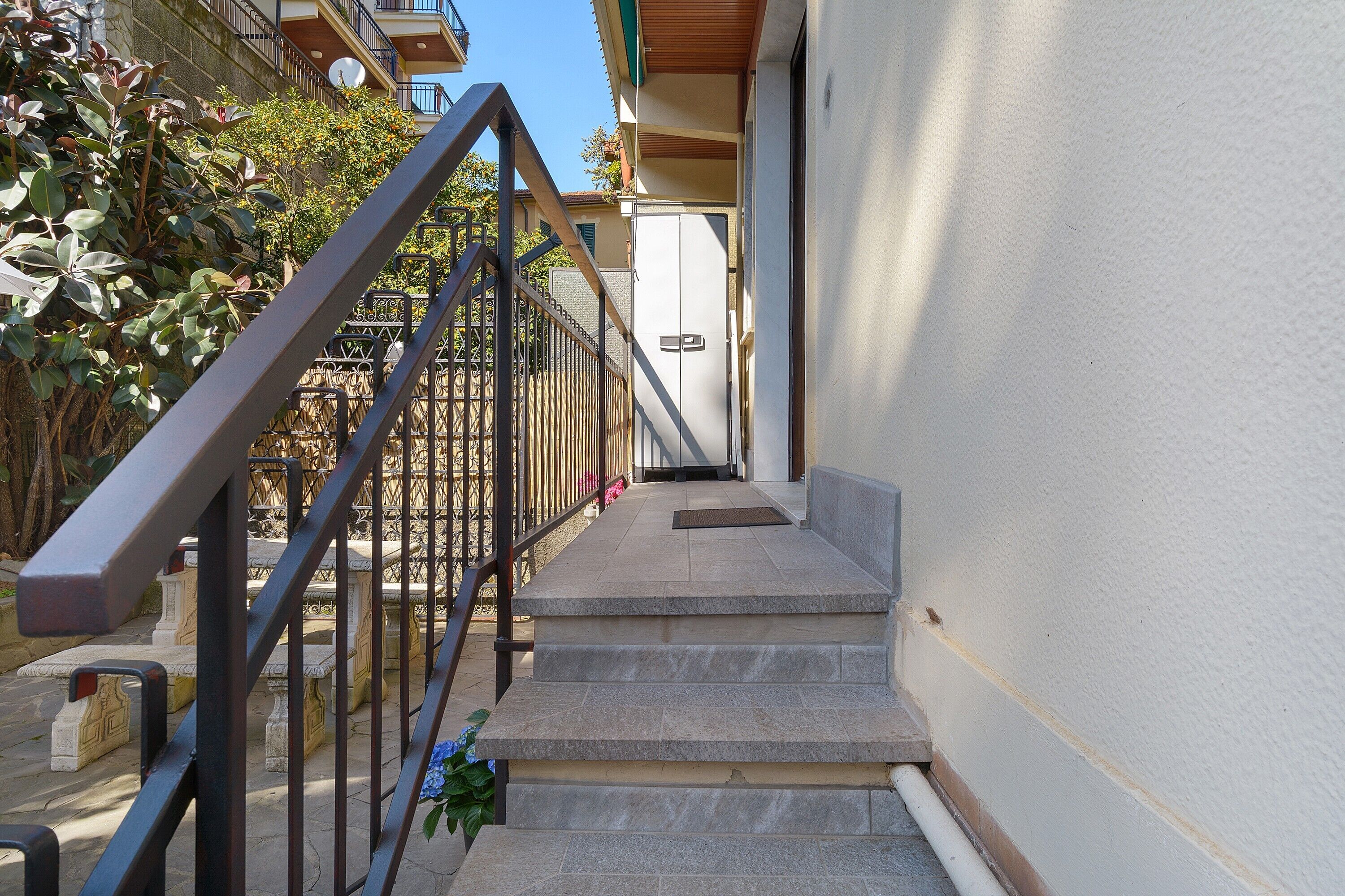Un escalier avec des rampes en métal menant à une porte, le tout entouré de plantes et adossé à la façade d’un immeuble.