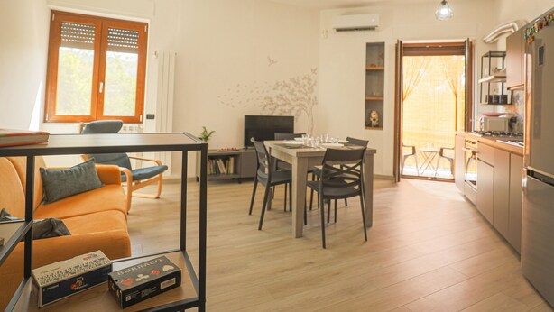 Un salon moderne comprenant un canapé, une table à manger et des chaises, un coin cuisine avec un réfrigérateur et une cuisinière, et une grande fenêtre dotée de volets en bois.