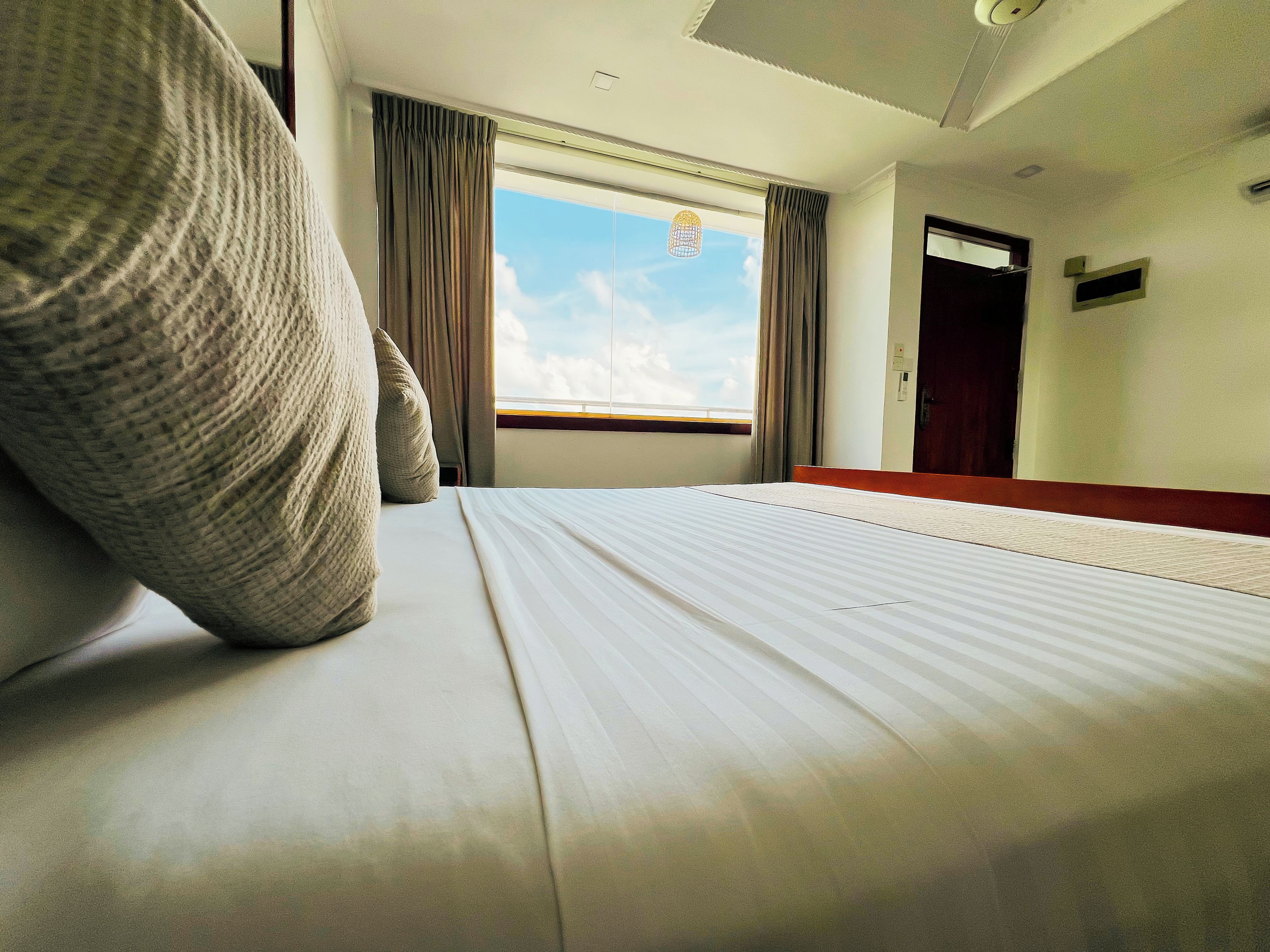 A hotel room with a bed, a window with curtains, a door, and a wall-mounted light fixture.