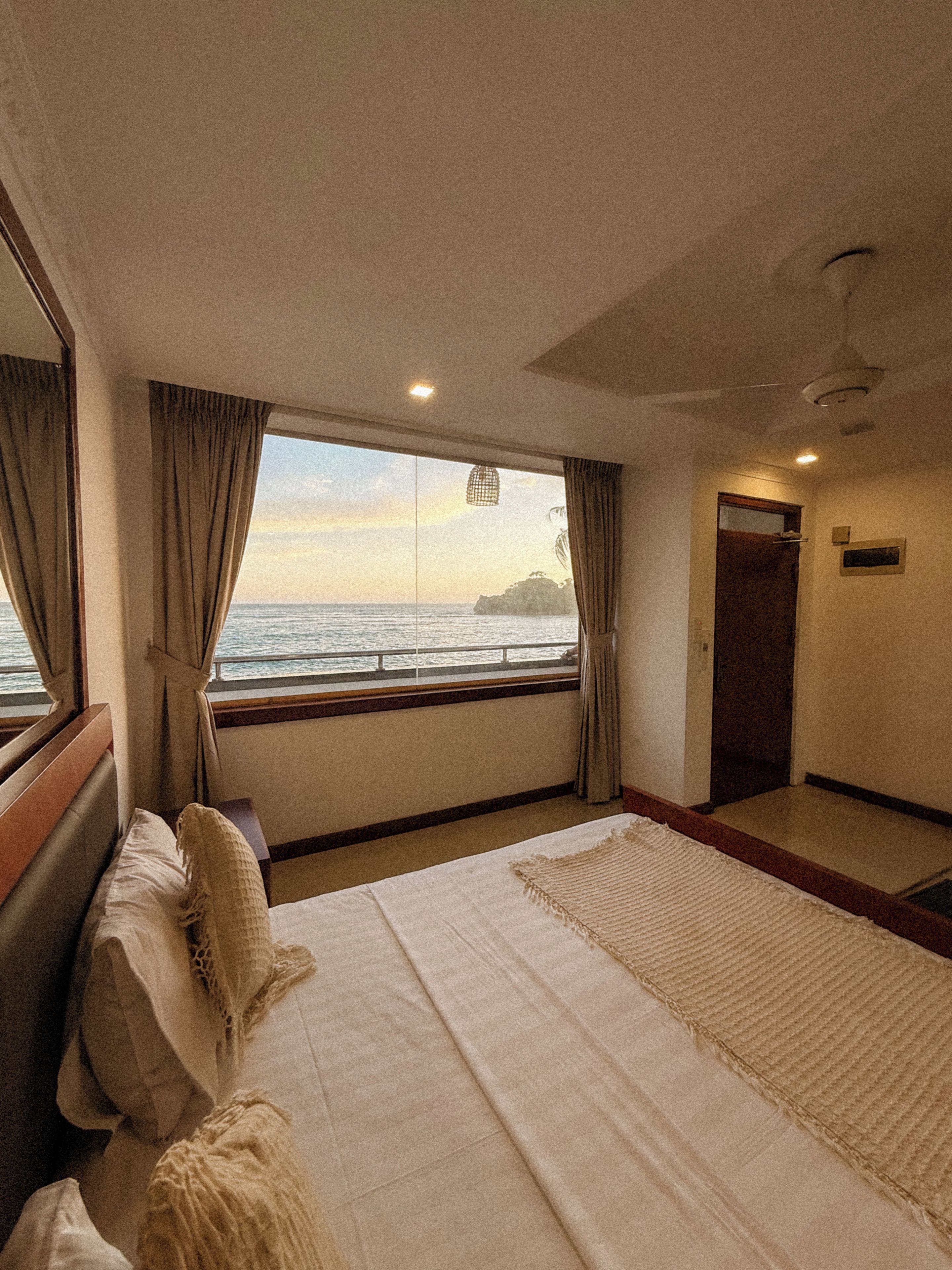 A bedroom with a large bed, a window with curtains, a view of the sea, and a ceiling fan.