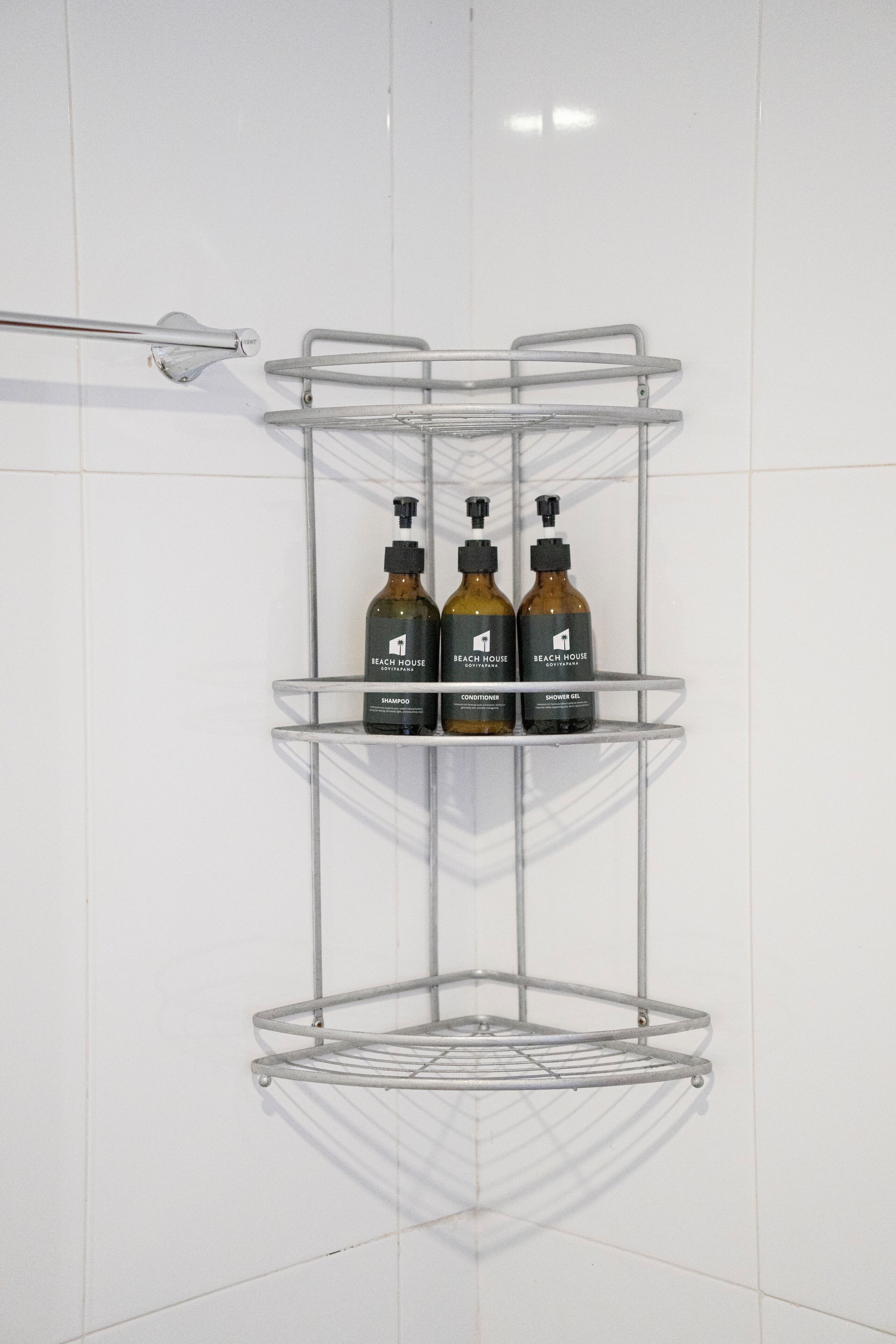A bathroom with a corner shelf unit holding three bottles of shampoo, conditioner, and shower gel.