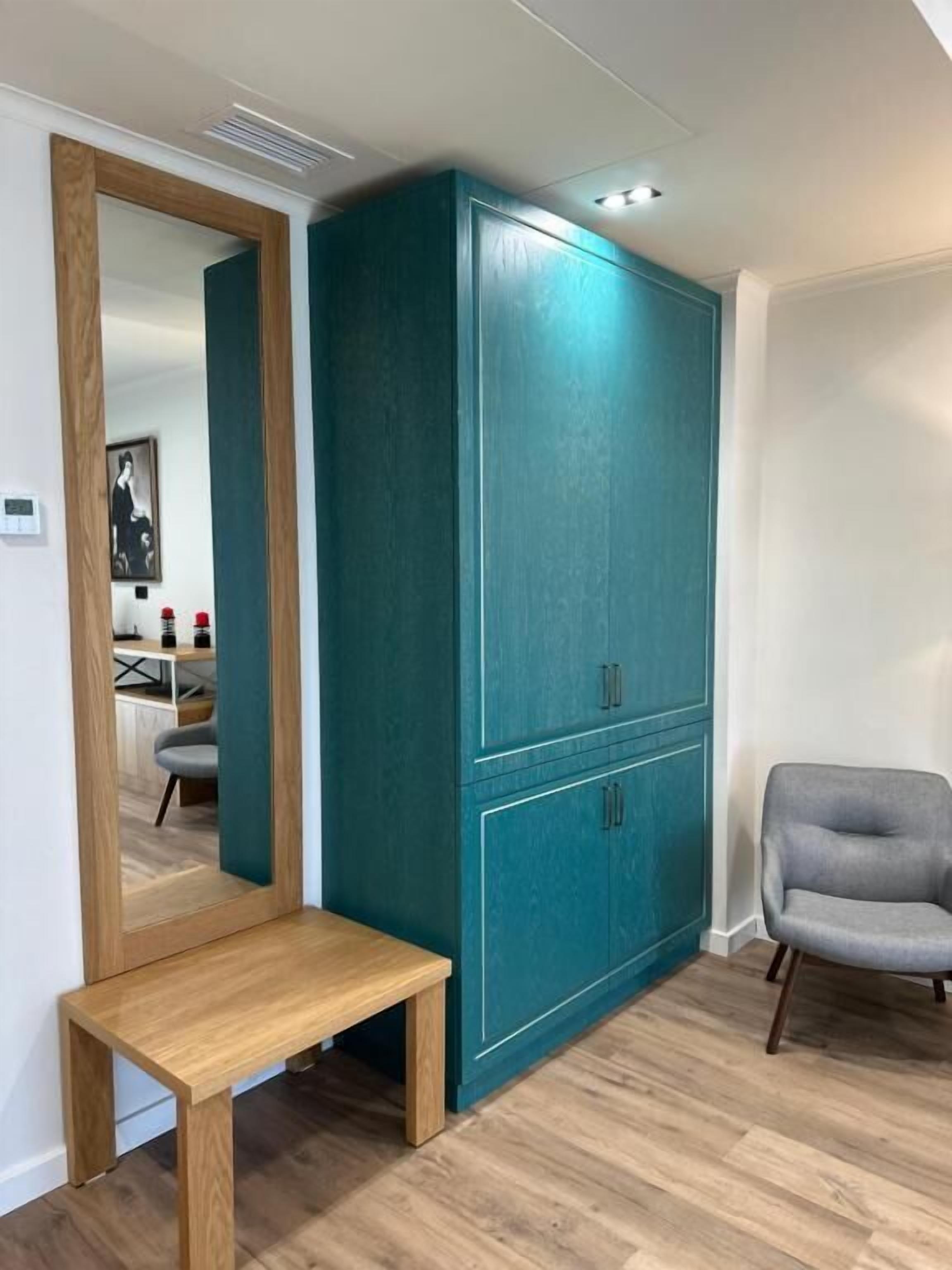 Una habitació amb un armari color turquesa, una banca de fusta i una poltrona gris.