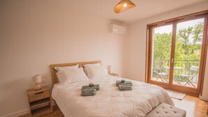 Un dormitorio con una cama grande, una mesita de noche de madera, un banco y vistas a un balcón con mobiliario exterior.