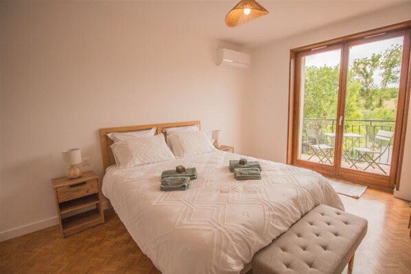 Un dormitorio con una cama grande, una mesita de noche de madera, un banco y vistas a un balcón con mobiliario exterior.