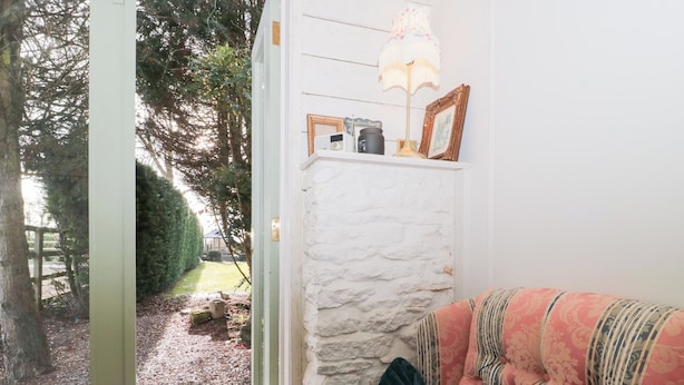 A room with a white brick wall, a wooden door, and a window. There is a patterned sofa with cushions and a lamp on a shelf. - Shades Farm Summerhouse (Swindon)