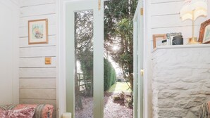 A cozy room with a wooden wall, a door leading to a garden, a cushioned bench, and a white stone fireplace. - Shades Farm Summerhouse (Swindon)