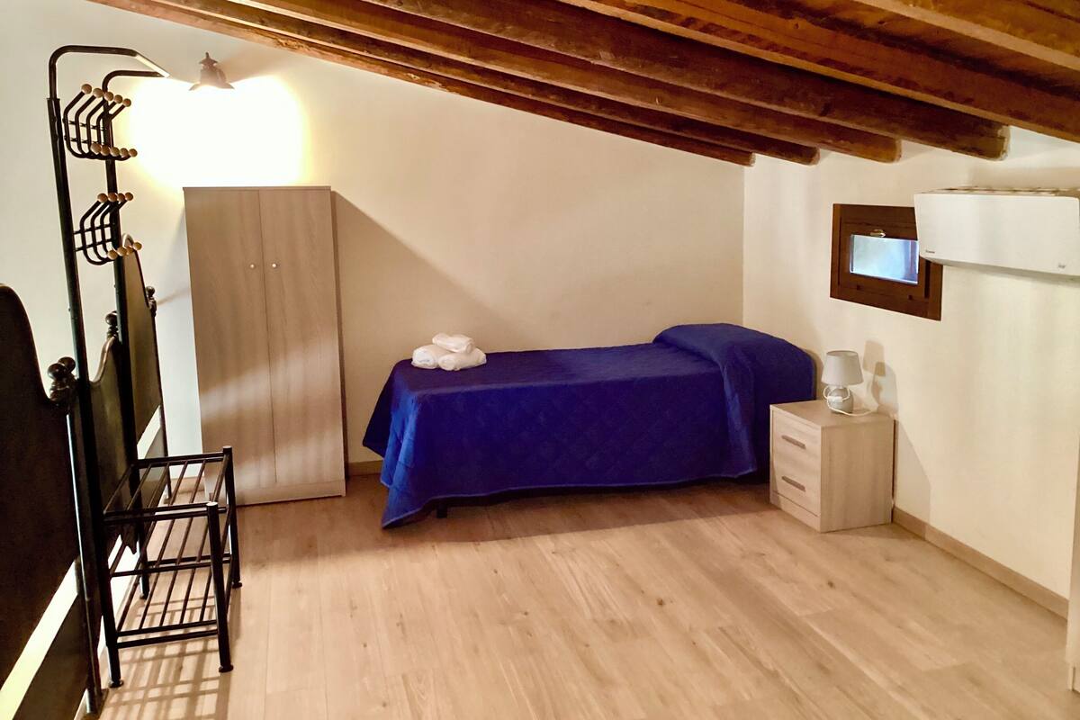 A small, neatly arranged bedroom with a bed, a wardrobe, a bedside table with a lamp, and a ceiling fan.