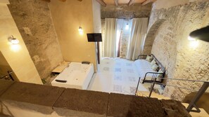 Una habitación con bañera, cama con colcha estampada, una mesita con lámpara y pared de piedra.