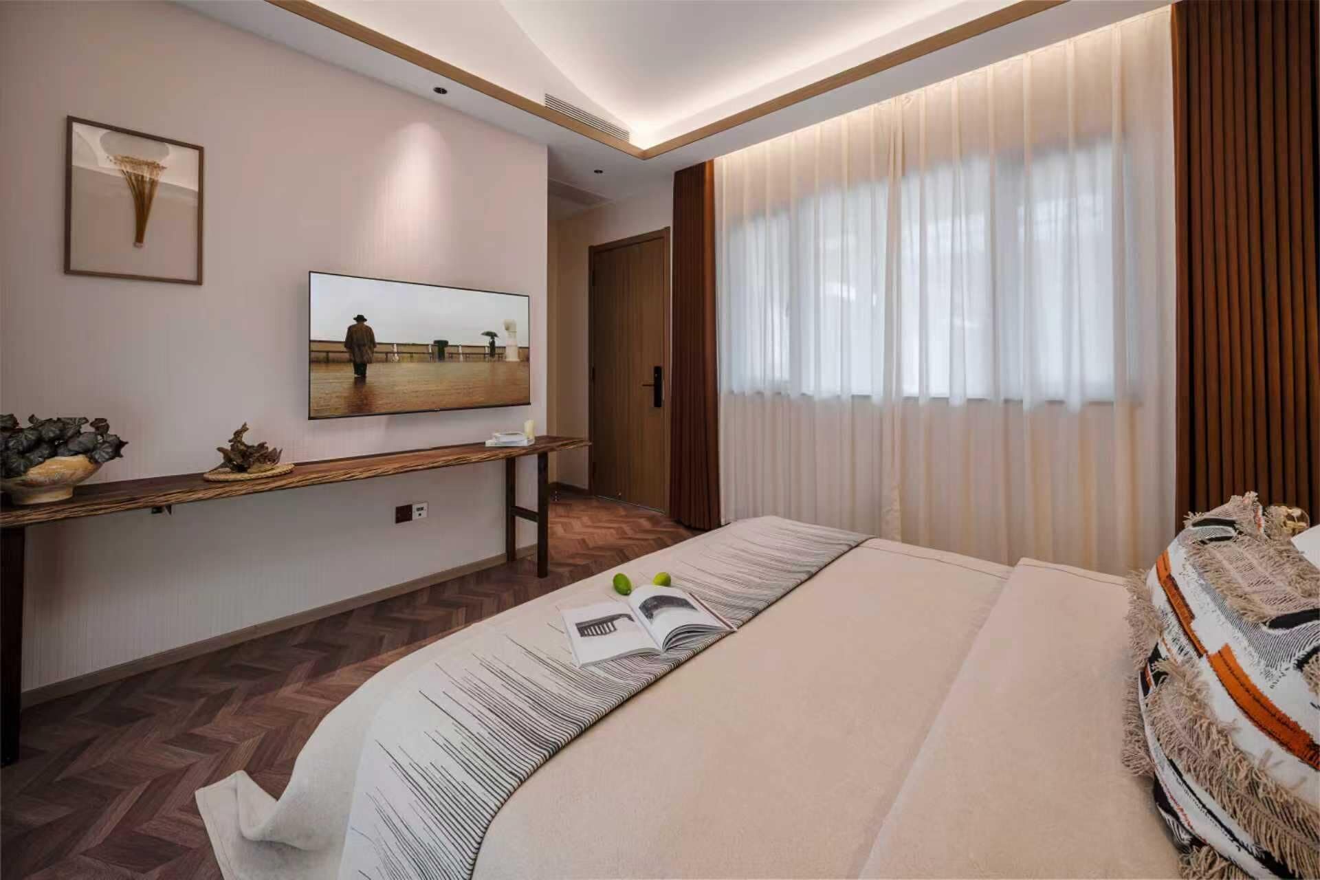 Quarto de hotel com uma cama, uma televisão sobre um console de madeira, uma janela grande com cortinas e um corredor que leva a outro quarto.