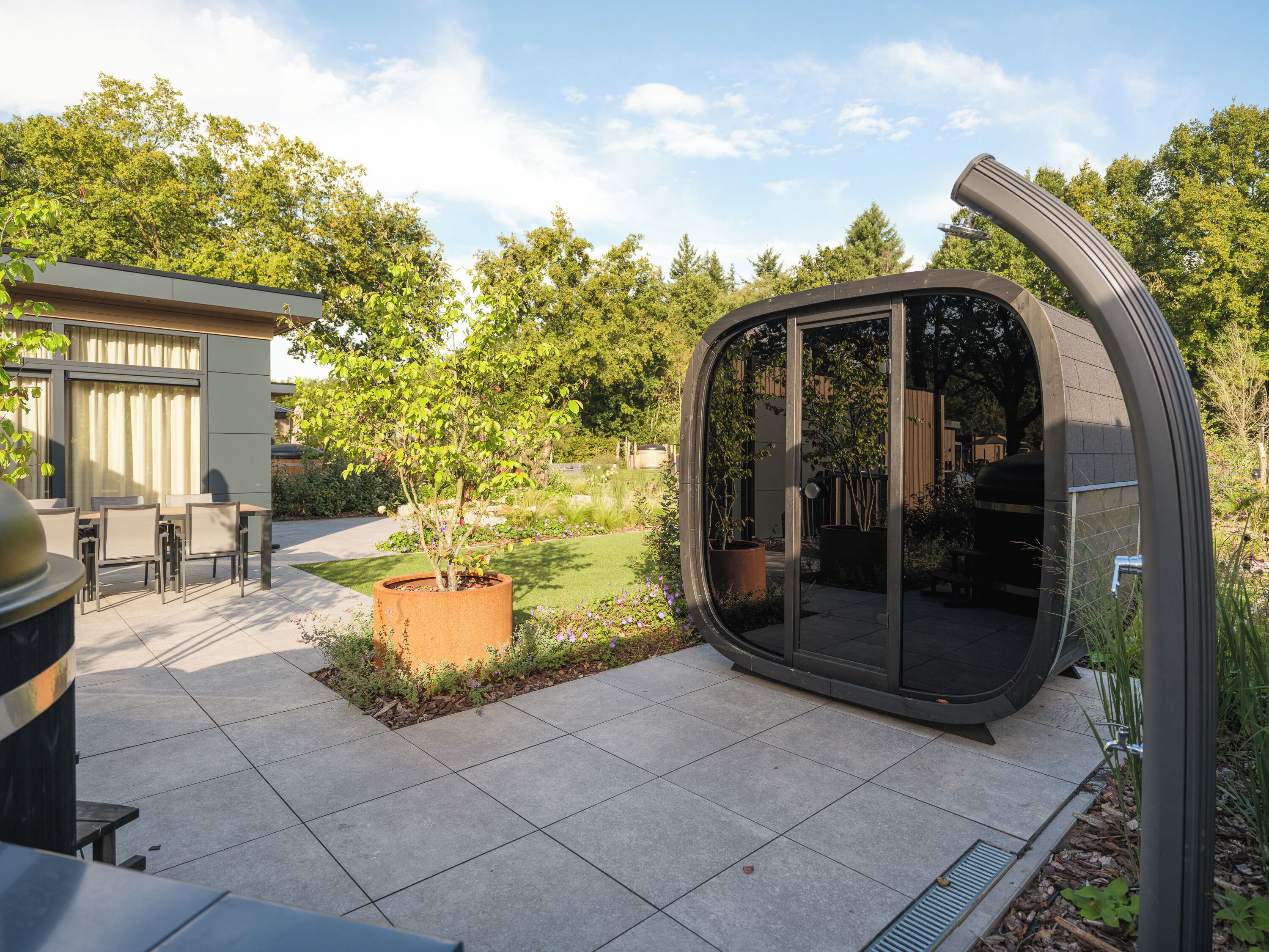 Eine moderne Terrassenanlage mit gewölbter Glasfassade, einem Essbereich mit Stühlen und einem kleinen Garten mit Baum und Blumen.
