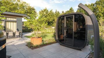 Eine moderne Terrassenanlage mit gewölbter Glasfassade, einem Essbereich mit Stühlen und einem kleinen Garten mit Baum und Blumen.