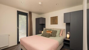 A bedroom with a bed, a bedside table with a lamp, a radiator, and a window with blinds. - Enjoy a private hot tub and sauna, a cosy outdoor lounge with fireplace and a luxurious outdoor area at the Cannenburch Deluxe. (Lieren)