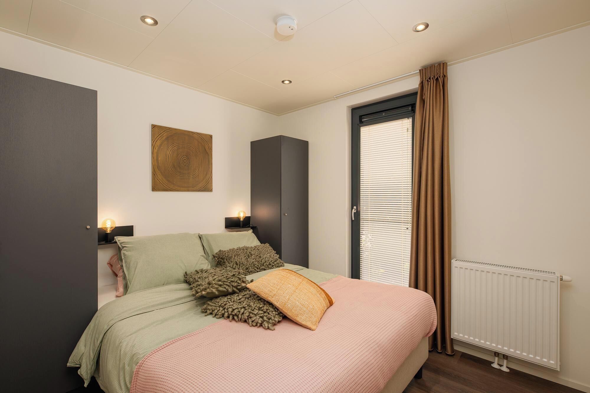 Une chambre à coucher avec un lit, des lampes de chevet, un radiateur et une fenêtre avec des rideaux.